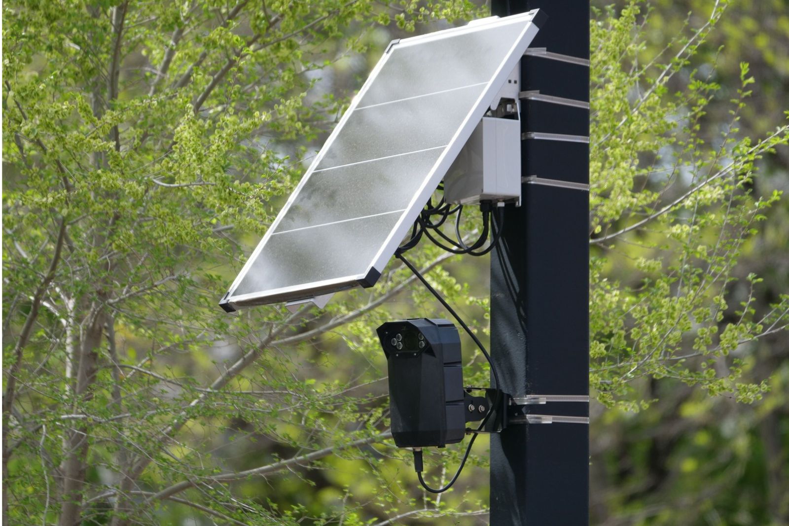A surveillance camera mounted beneath a solar panel on a pole, surrounded by green trees.