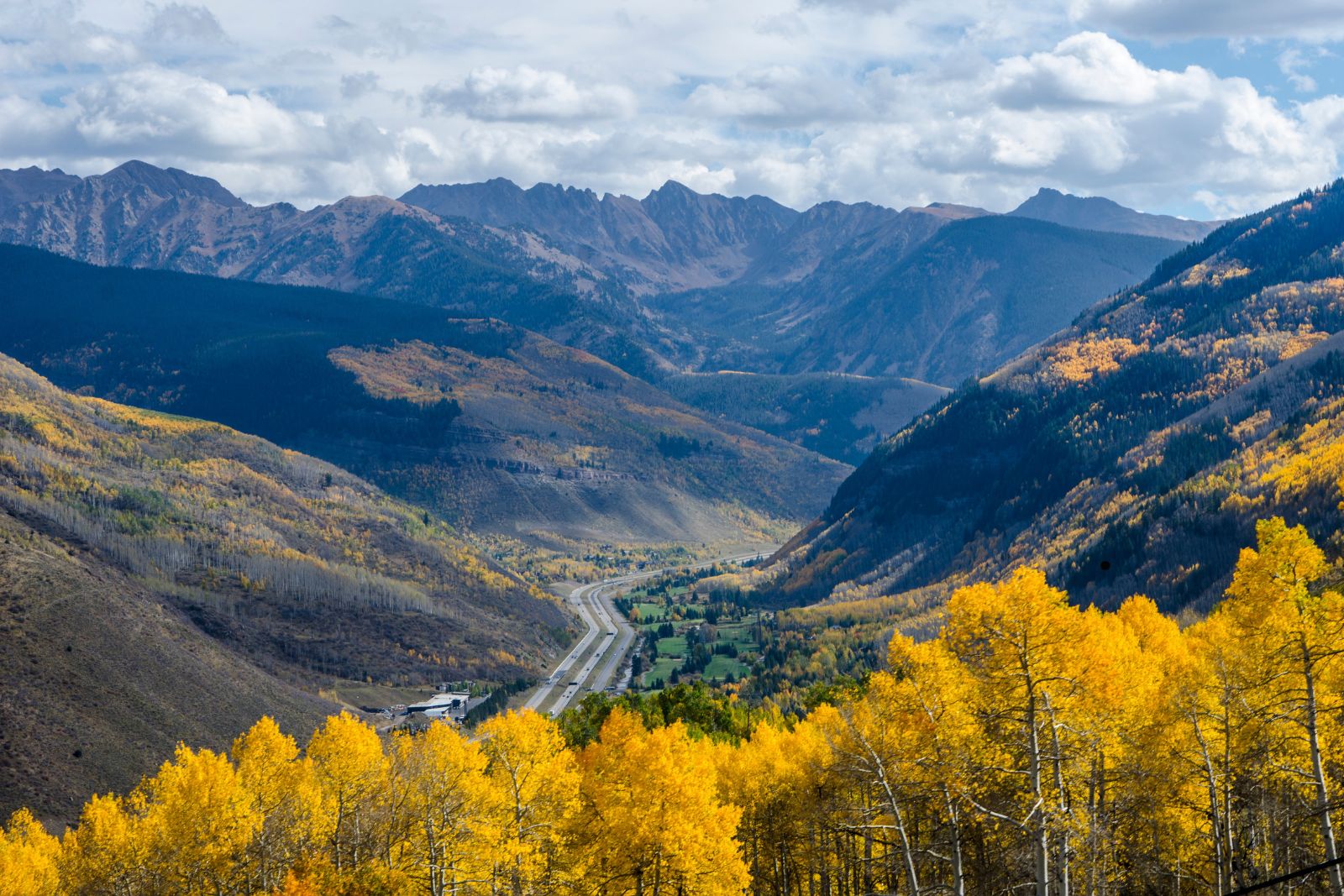 A divided highway winds through a mountain valley surrounded by golden aspens and forested slopes.