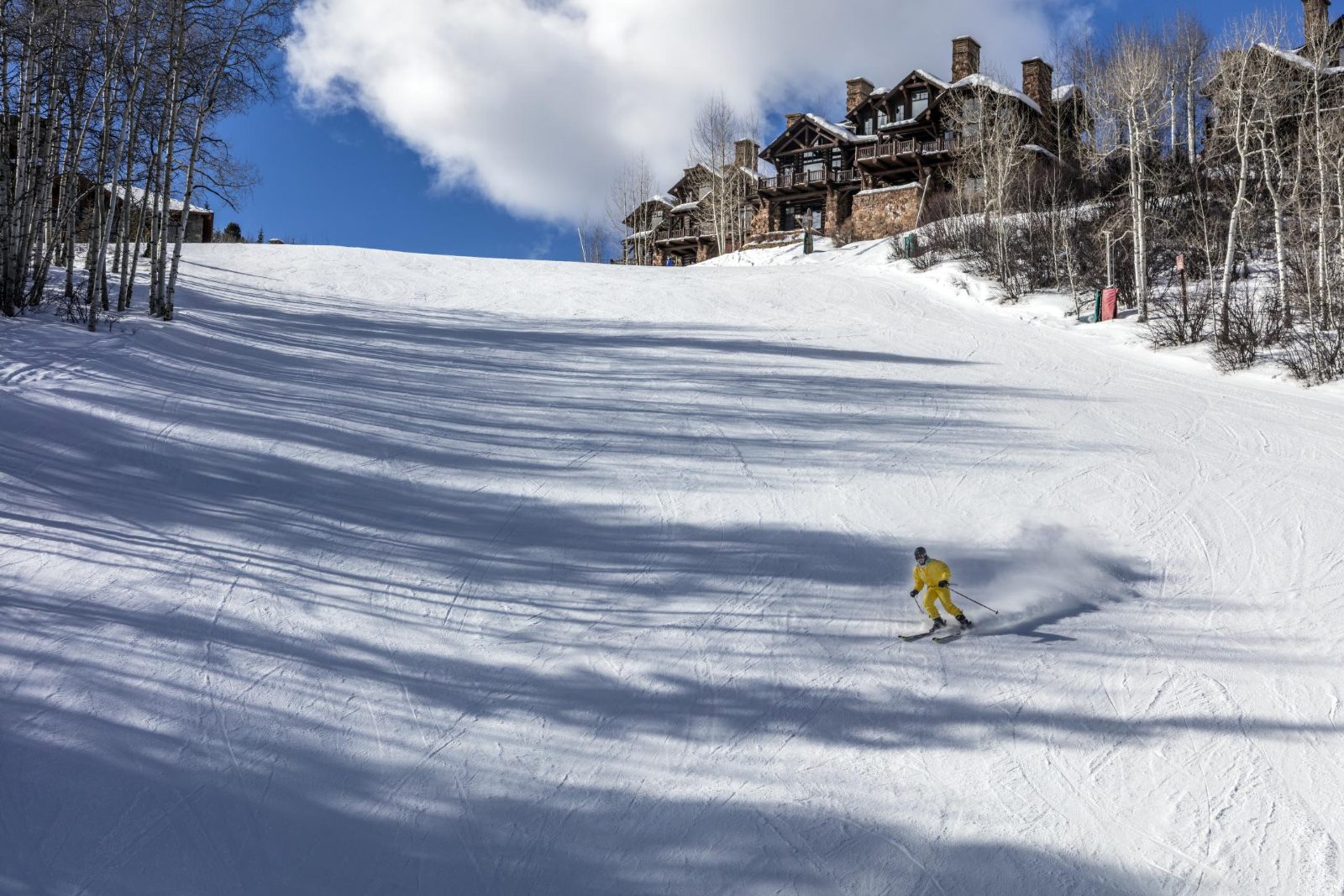 A skier in a yellow suit descends a groomed run in Bachelor Gulch, with aspen trees and slopeside homes at the top of the slope
