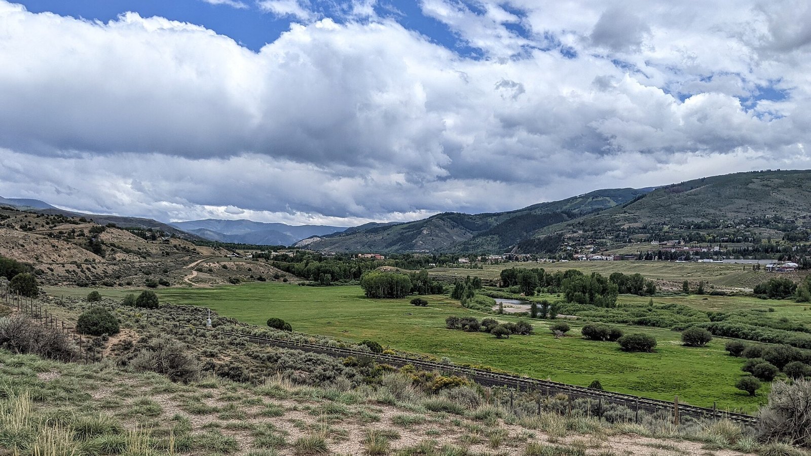 Green ranch meadows and sagebrush slopes in the Eagle River valley under a cloudy summer sky, with Edwards in the distance.