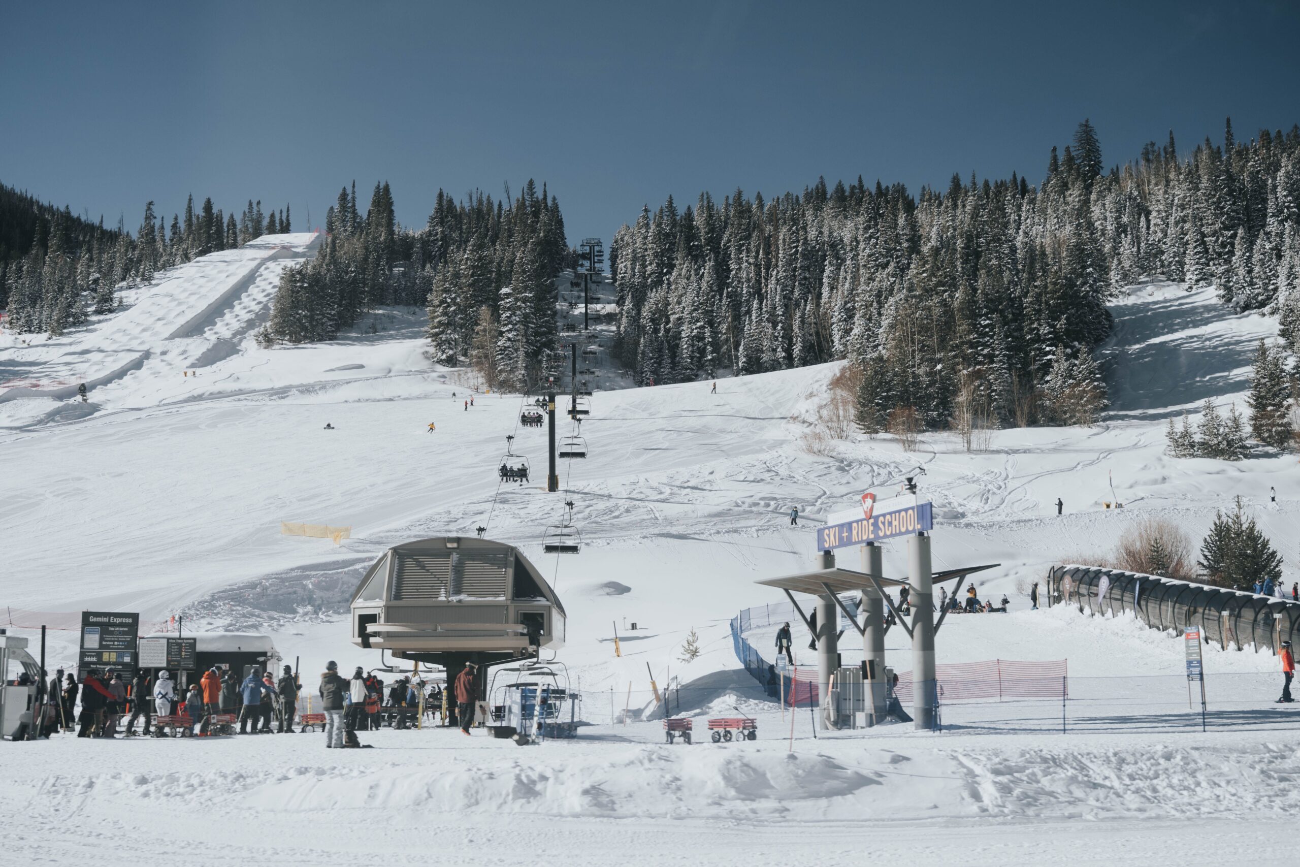 Skiers gather beside a chairlift below snow-covered slopes and evergreen trees.