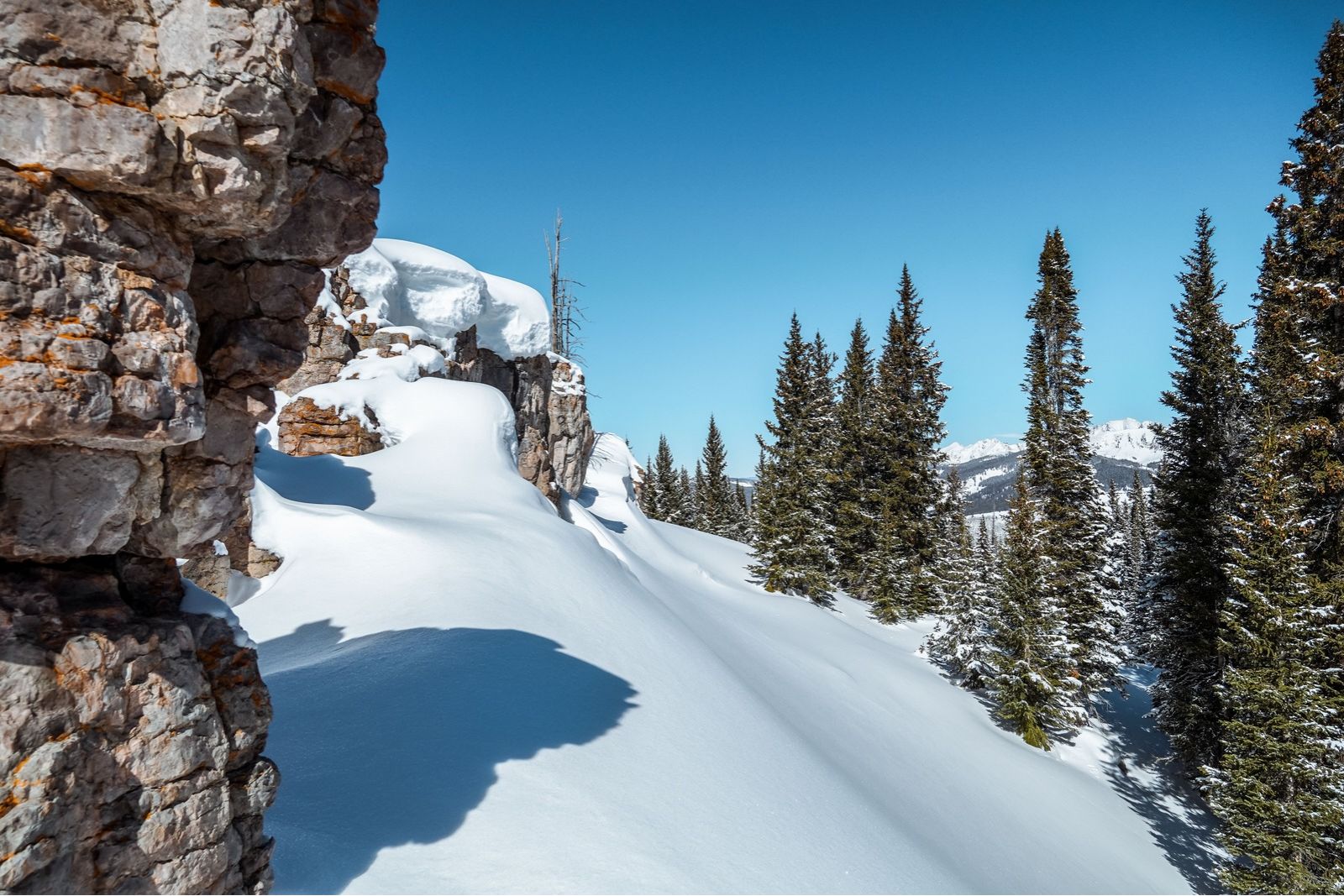 Snow-covered rock ledges and evergreen trees beneath a clear blue sky in Vail, Colorado.
